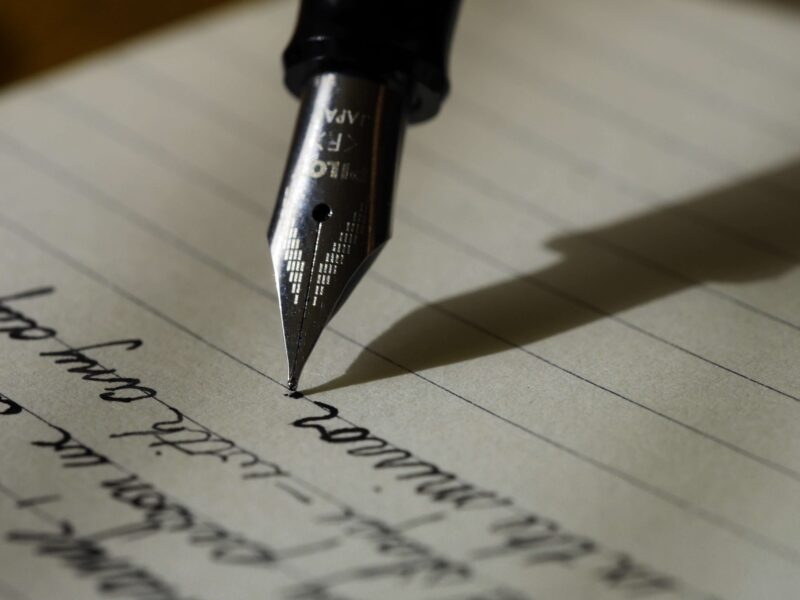 Close-up of a fountain pen writing in cursive on lined paper. The pen's nib is clearly visible, casting a shadow on the paper. The handwriting is in dark ink, partially showing some written words.