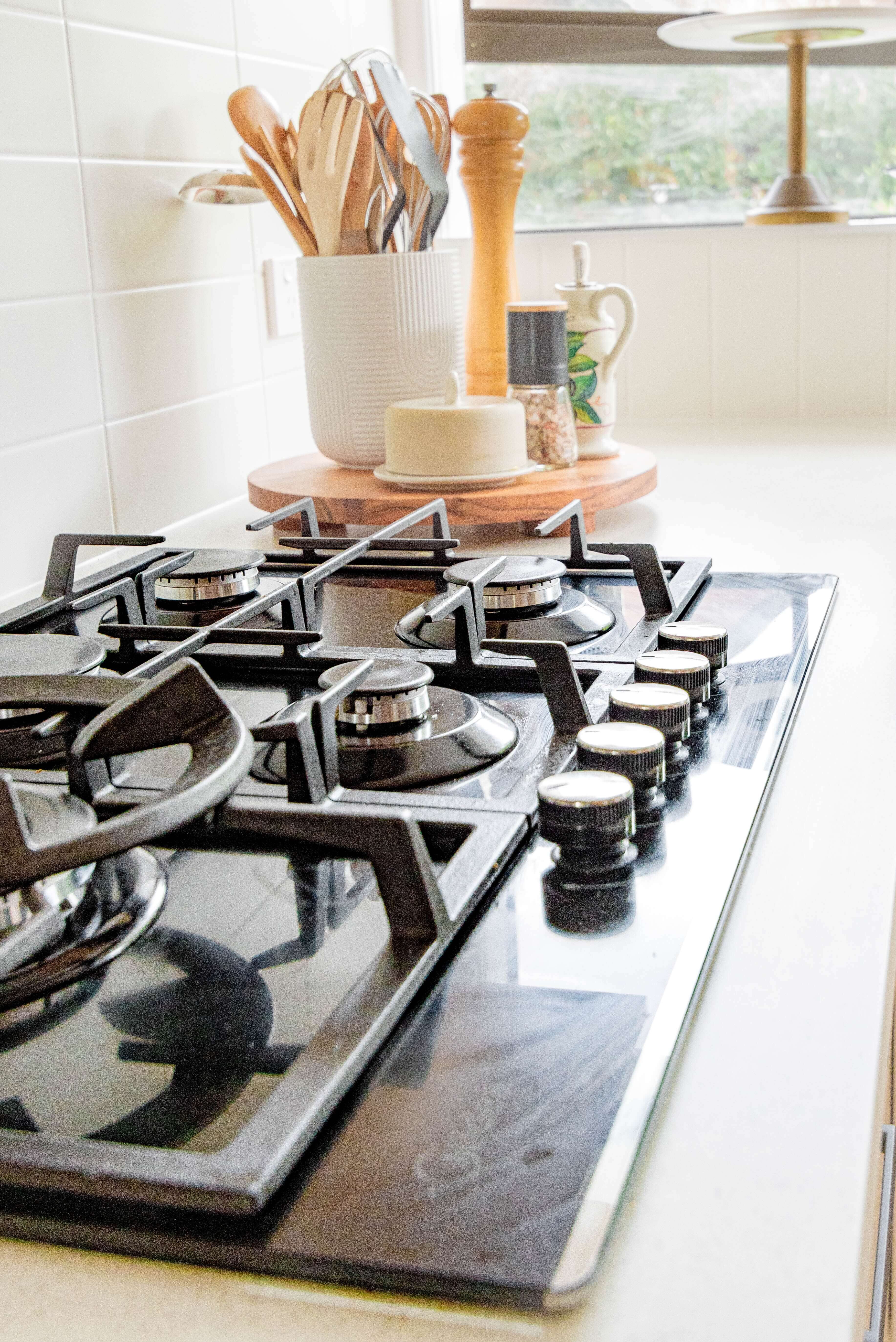 Close-up of a modern kitchen stove with black gas burners and control knobs. A nearby countertop holds a utensil holder with wooden spoons, a pepper grinder, a butter dish, and small potted herbs by a window.