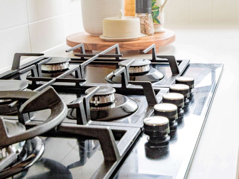 Close-up of a modern kitchen stove with black gas burners and control knobs. A nearby countertop holds a utensil holder with wooden spoons, a pepper grinder, a butter dish, and small potted herbs by a window.