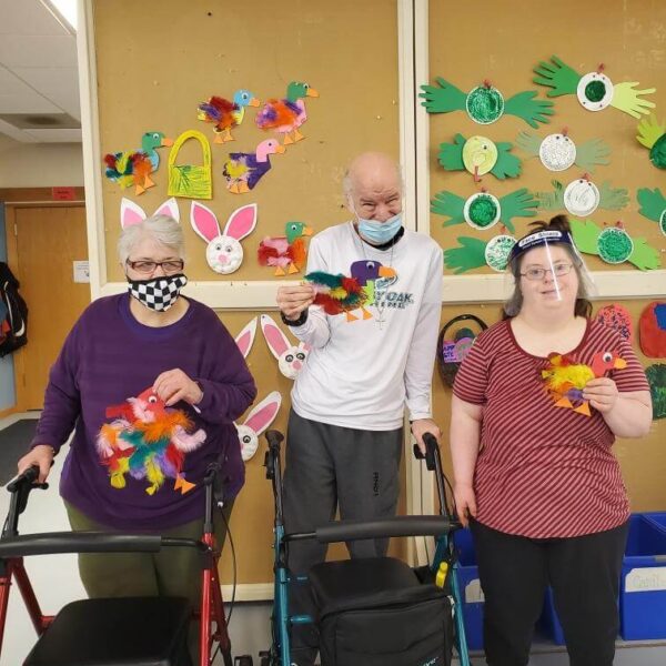 Three people smile while holding colorful bird crafts. They stand in front of a wall decorated with paper bunnies and birds. Two use walkers, and one wears bunny ears. The atmosphere is cheerful and creative.