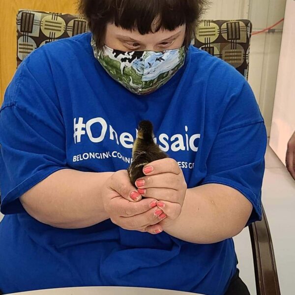 A person wearing a blue "#OneMosaic" shirt and a floral mask is gently holding a small duckling in their hands while sitting at a table with a coloring page.