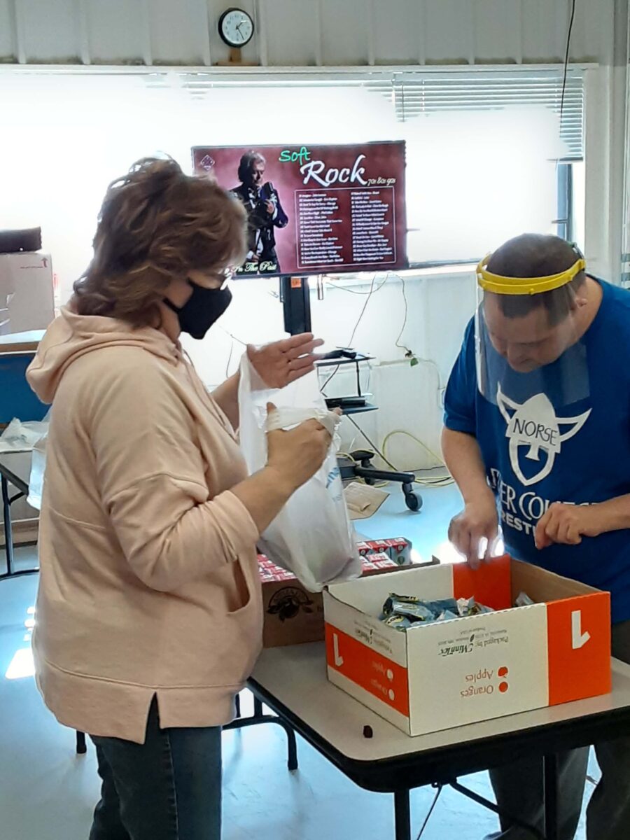 Two people wearing face masks and shields work together at a table with boxes of supplies. One person holds a plastic bag while the other sorts items. A screen displaying 