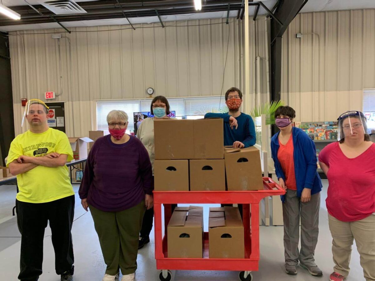 A group of people wearing face masks and standing indoors around a red cart filled with cardboard boxes. They are in a spacious, industrial-style building with visible ceiling beams and fluorescent lights.