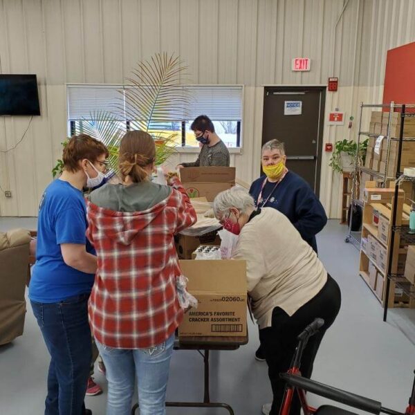 People are gathered around tables in a room, organizing items for a food bank. They are wearing masks and gloves. Shelves filled with boxes are visible in the background.