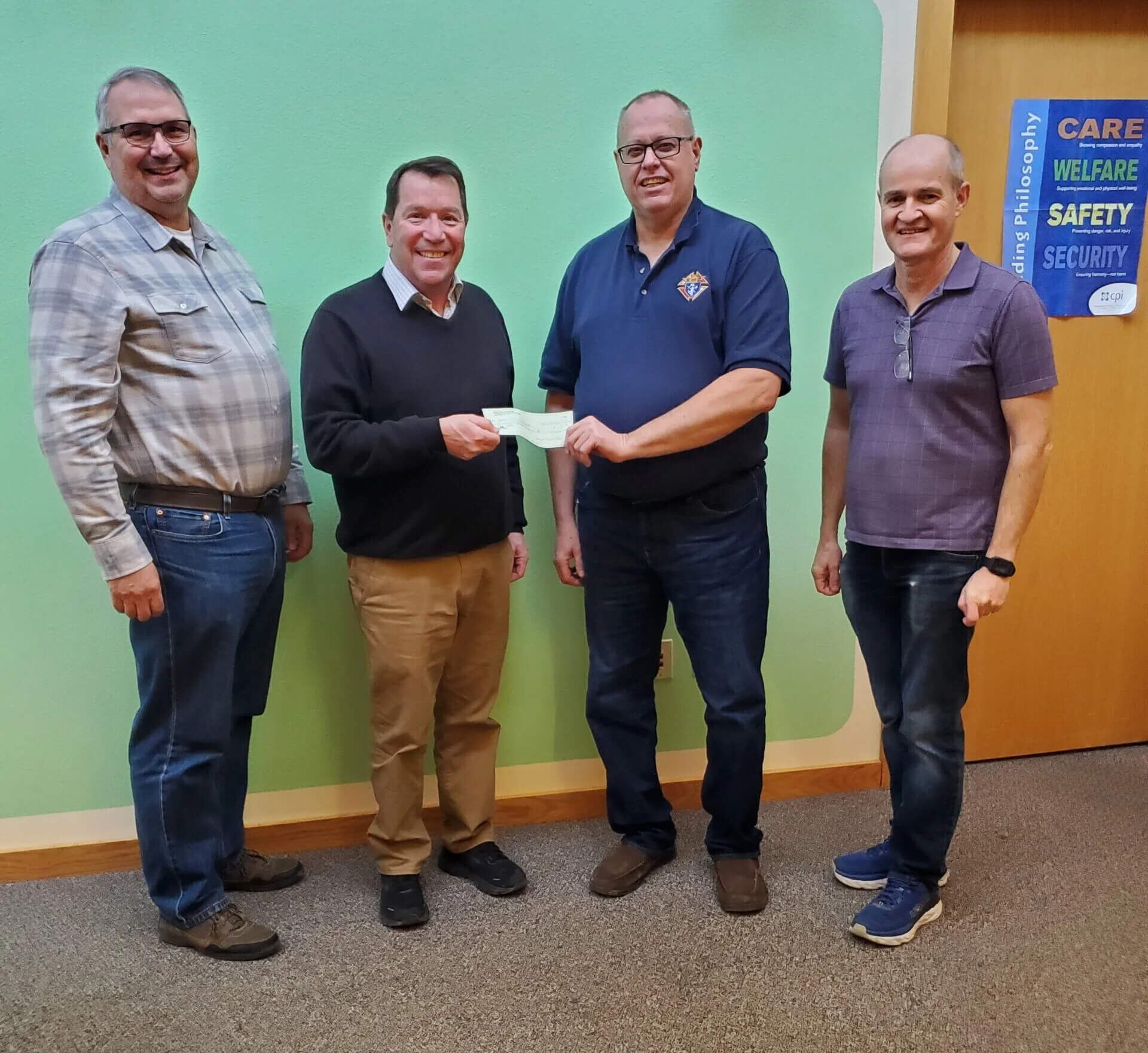 Four men are standing indoors on a carpeted floor. Two of them are holding a check between them. They are casually dressed, and there is a green wall with signs about care, welfare, safety, and security in the background.