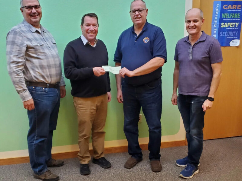 Four men are standing indoors on a carpeted floor. Two of them are holding a check between them. They are casually dressed, and there is a green wall with signs about care, welfare, safety, and security in the background.