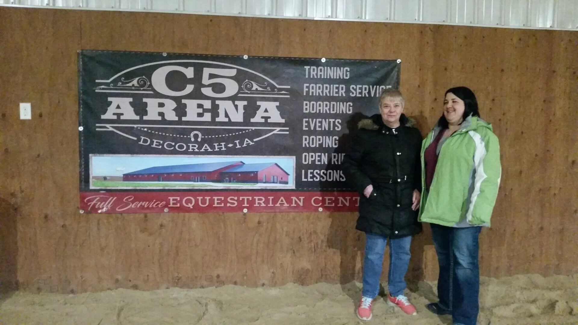 Two people stand in front of a sign for C5 Arena in Decorah, IA. The sign mentions services like training, farrier service, boarding, roping, and events, indicating it's a full-service equestrian center. The setting appears to be an indoor arena.