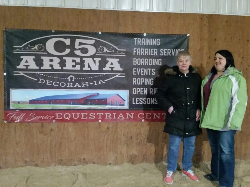 Two people stand in front of a sign for C5 Arena in Decorah, IA. The sign mentions services like training, farrier service, boarding, roping, and events, indicating it's a full-service equestrian center. The setting appears to be an indoor arena.