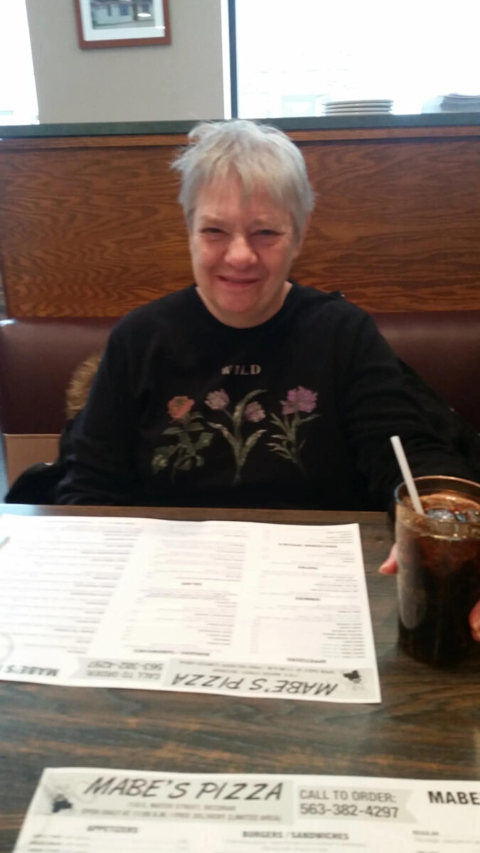 An older woman with short gray hair is sitting in a restaurant booth, smiling at the camera. She is wearing a black sweatshirt with floral designs and has a drink with a straw on the table in front of her, along with a menu.