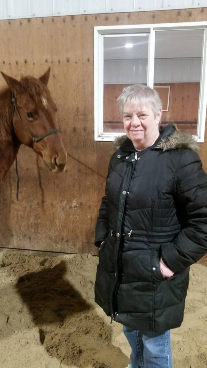 A person in a black coat stands inside a stable next to a brown horse. The person is smiling, with their hand in their pocket, and the stable wall has a window behind them.