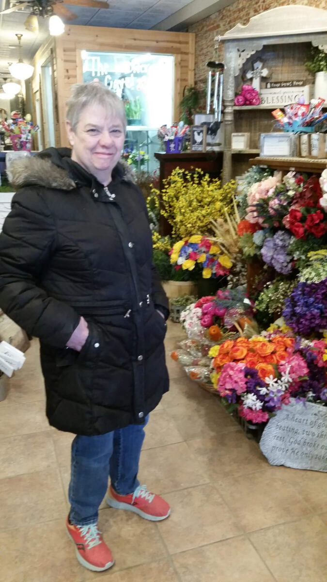 A person in a black coat stands smiling inside a flower shop, surrounded by colorful bouquets of flowers including roses and hydrangeas. The shelves in the background display various floral arrangements and decorations.