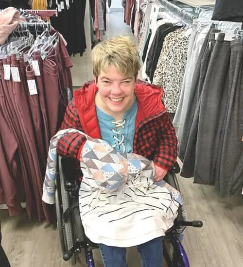 A person in a wheelchair smiles while holding clothing in a store. They wear a red plaid jacket and a blue shirt, surrounded by various clothes on racks.