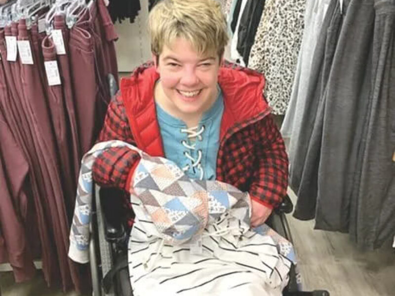 A person in a wheelchair smiles while holding clothing in a store. They wear a red plaid jacket and a blue shirt, surrounded by various clothes on racks.