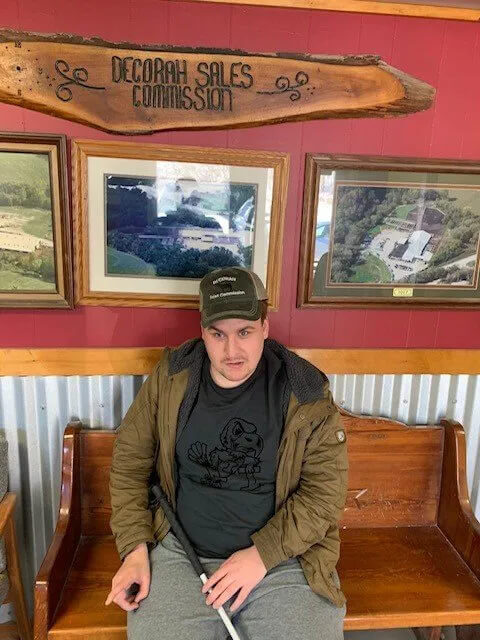 A person sitting on a wooden bench in front of a red wall with framed pictures. The bench is below a wooden sign that reads 