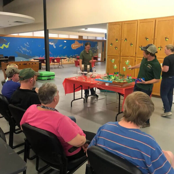 A group of people sits listening to a presentation in a room. Two presenters stand behind a table covered with models and educational materials. The walls are decorated with colorful designs.