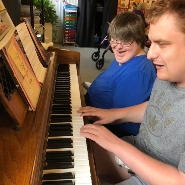 Two people sit side by side at a piano, playing music together. The person on the left is wearing a blue shirt, and the person on the right is wearing a gray shirt. There are colorful decorations and a walker in the background.