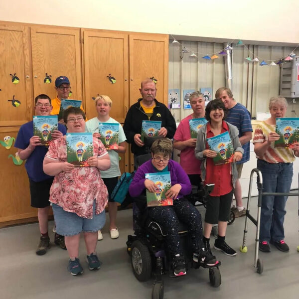 A group of people smiling and holding colorful children's books. They are standing in a classroom setting with wooden cabinets and paper decorations on the wall. One person is seated in a wheelchair, and another uses a walker.