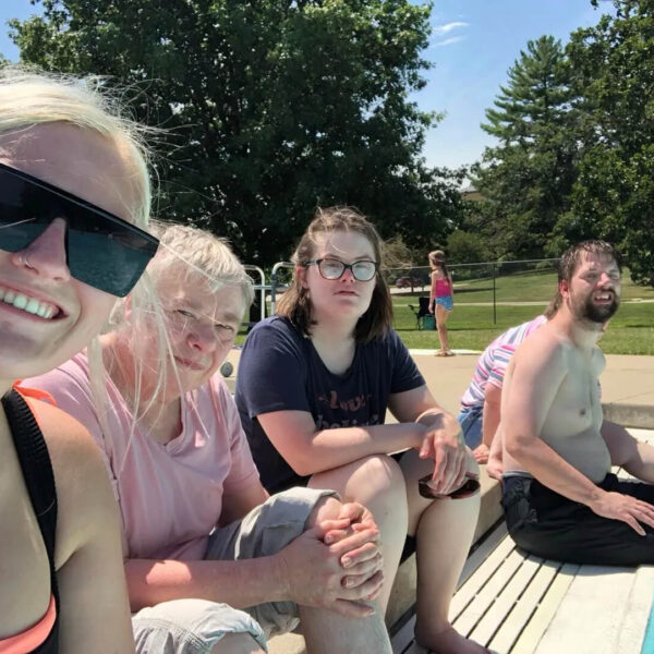 A group of people sitting by a pool, with some dipping their feet in the water. They are outdoors, enjoying a sunny day. There are trees and a grassy area in the background.