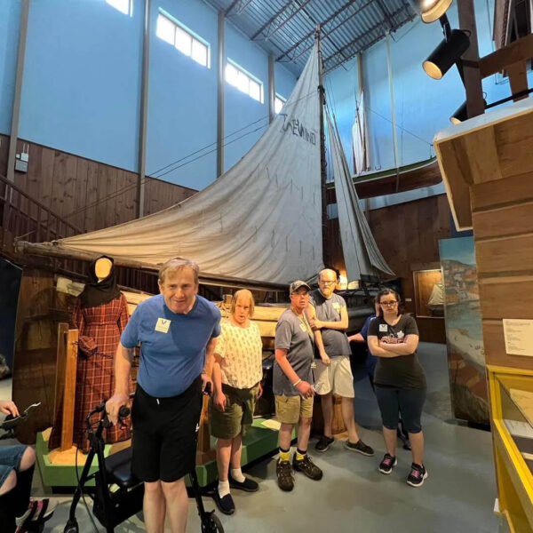 A group of people poses in a museum in front of a large, historic wooden boat with a white sail. The room features high ceilings and wooden walls, creating a nautical atmosphere.