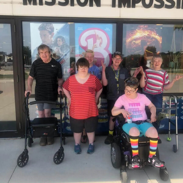 A group of five people smiling in front of a movie theater entrance with "Mission Impossible" on the marquee. Two people have walkers, and one is in a wheelchair. They are wearing casual summer clothing.