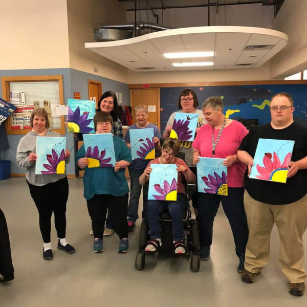 A group of people holding up colorful paintings featuring purple flowers and a sun. One person is seated in a wheelchair. They are standing in a room with art supplies visible in the background.