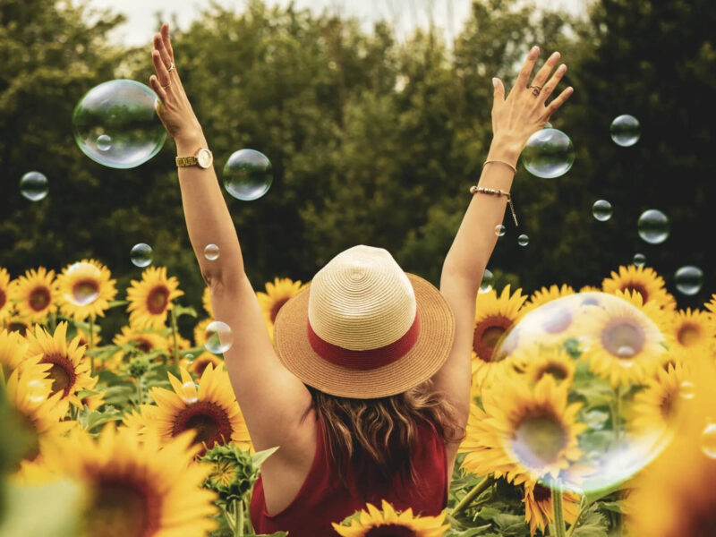 A person wearing a straw hat raises their arms joyfully in a field of sunflowers. Bubbles float around them, and green trees are in the background.