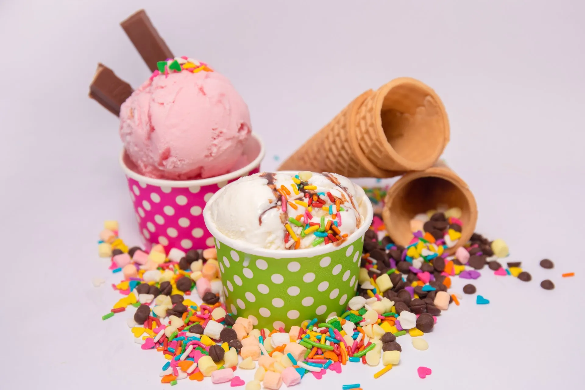 Two ice cream scoops in colorful polka dot cups, one pink and one white with sprinkles, surrounded by colorful sprinkles, chocolate pieces, and two empty waffle cones on a light background.