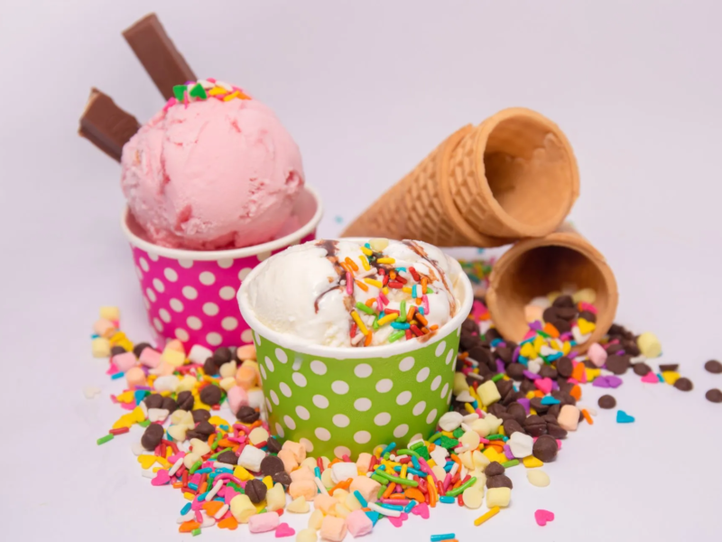 Two ice cream scoops in colorful polka dot cups, one pink and one white with sprinkles, surrounded by colorful sprinkles, chocolate pieces, and two empty waffle cones on a light background.