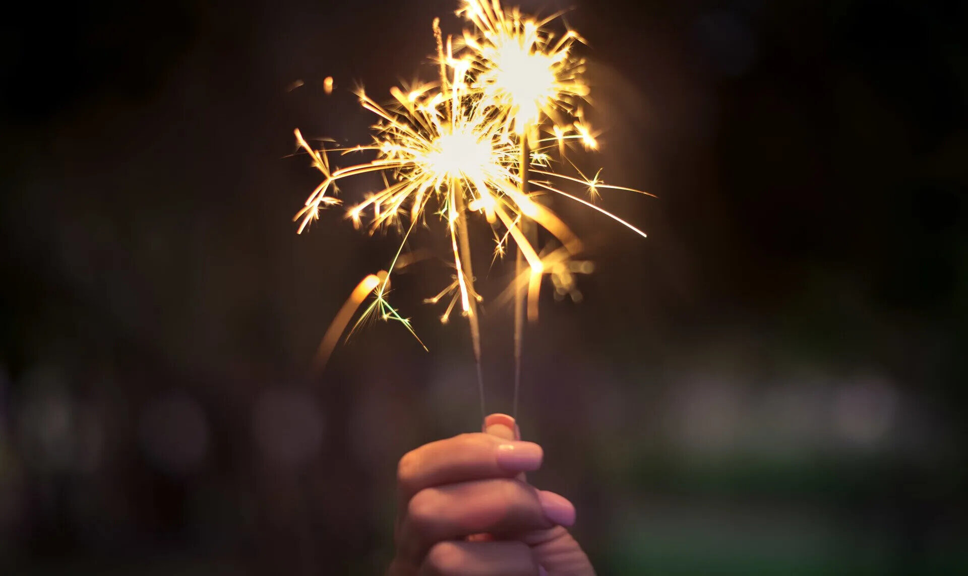 A hand holding two lit sparklers against a dark background. The sparklers emit bright, glowing sparks and streaks of light, creating a festive and lively atmosphere.