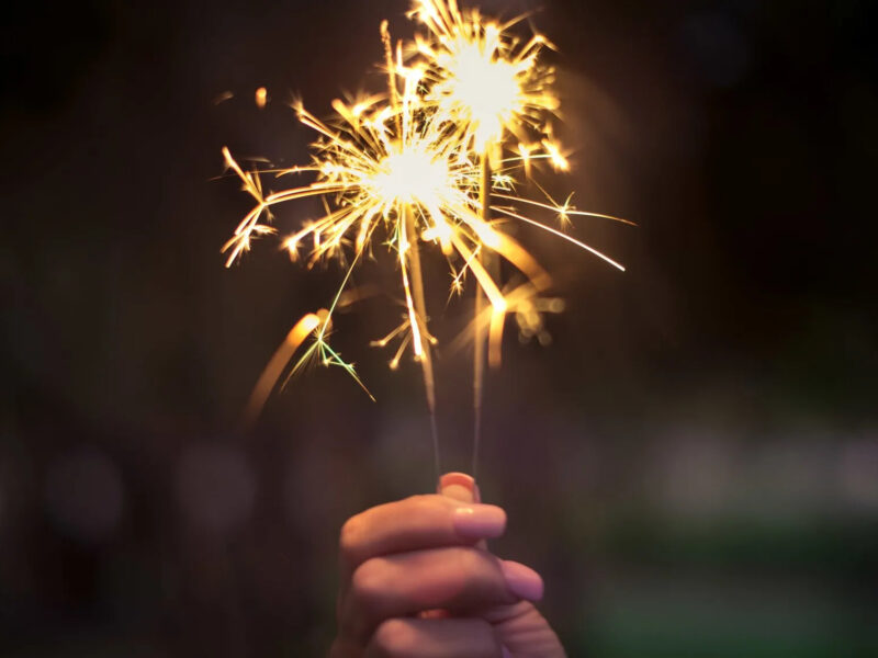 A hand holding two lit sparklers against a dark background. The sparklers emit bright, glowing sparks and streaks of light, creating a festive and lively atmosphere.