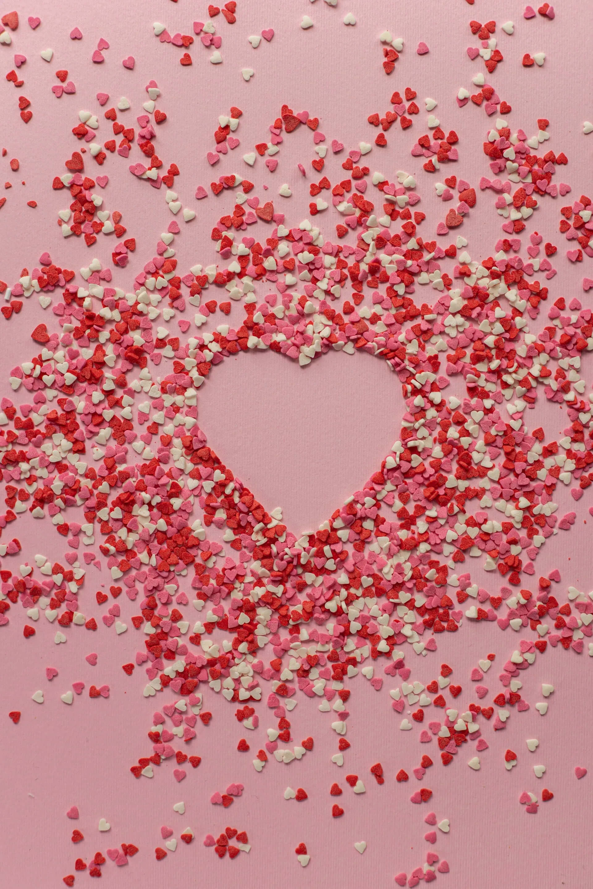 A heart shape is formed on a pink background, surrounded by scattered small heart-shaped confetti in shades of red, pink, and white.