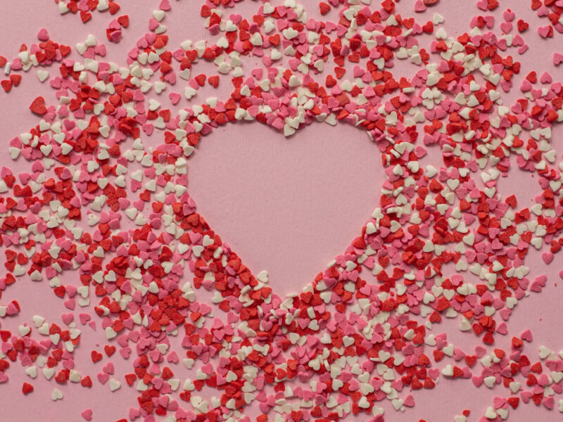 A heart shape is formed on a pink background, surrounded by scattered small heart-shaped confetti in shades of red, pink, and white.