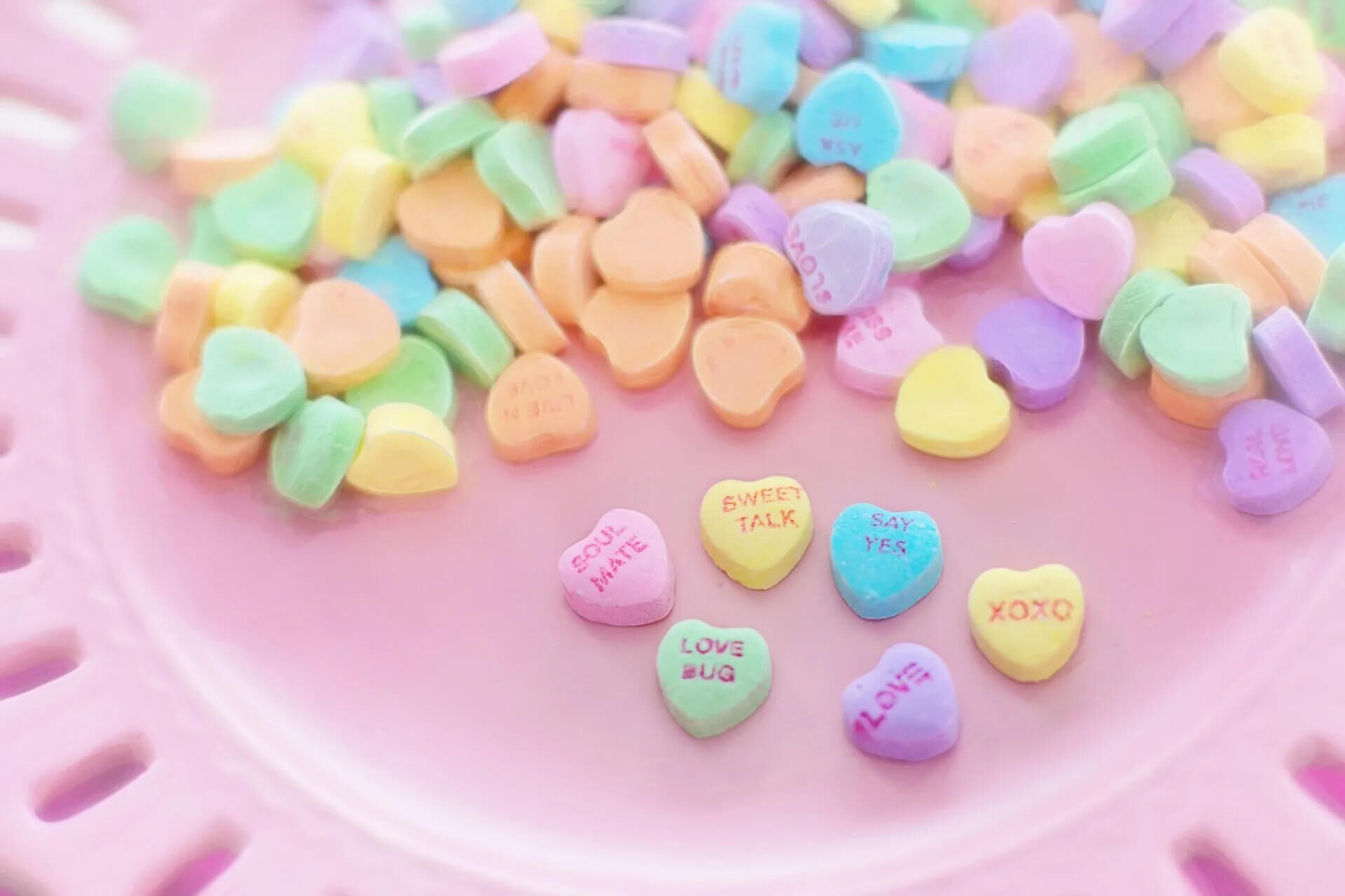 A collection of pastel-colored heart-shaped candies on a pink surface, some with words like 