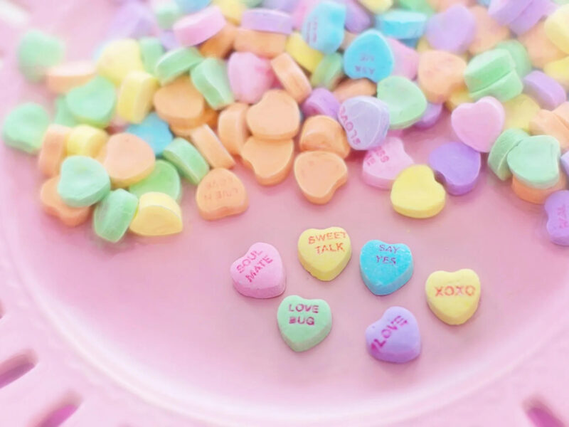 A collection of pastel-colored heart-shaped candies on a pink surface, some with words like 